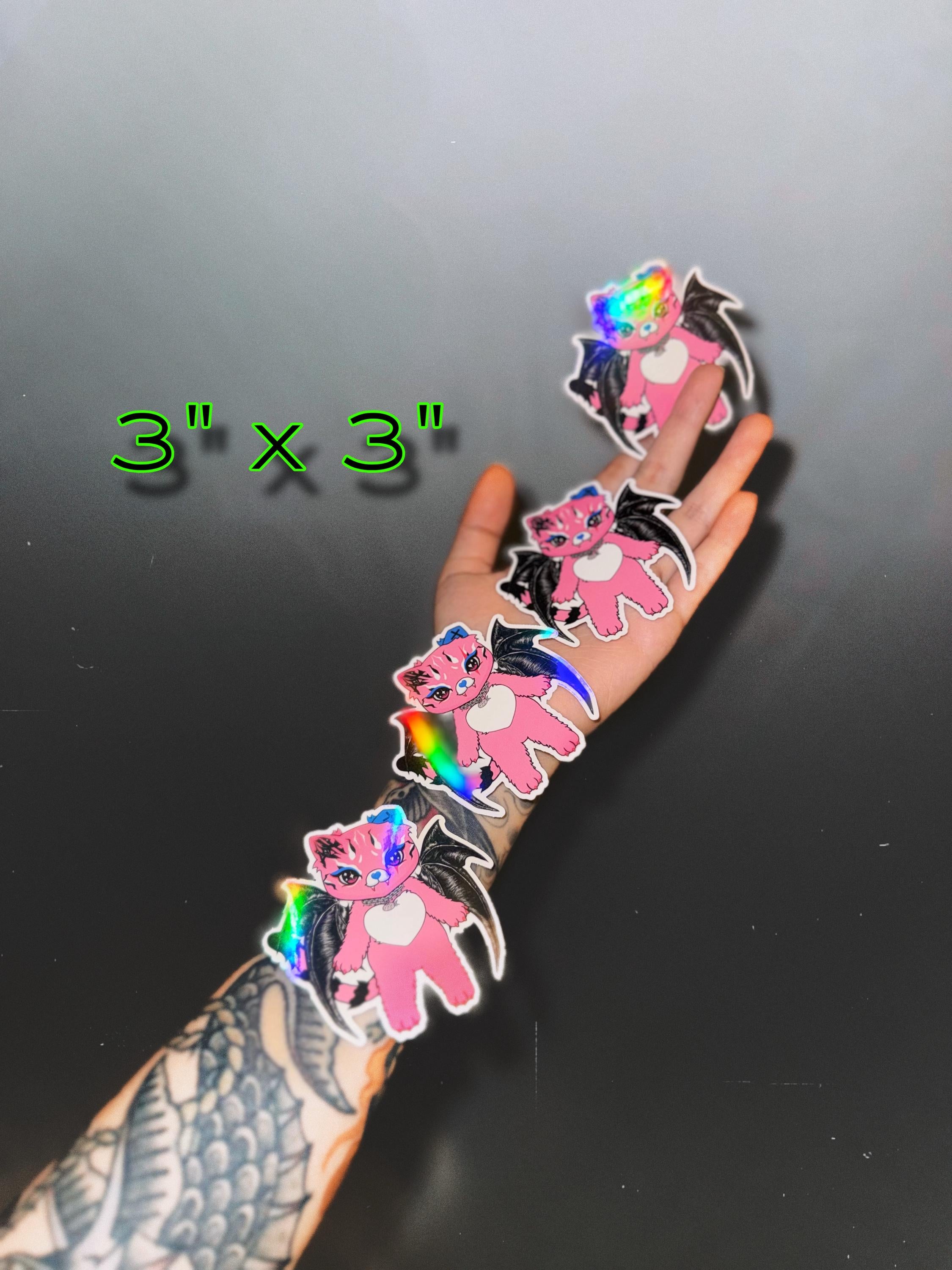 Tommi the Batcat holographic sticker being held in hand with stickers placed on forearm, showing actual 3 x 3 inch size for comparison reference